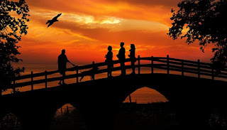 Uma pessoa com cão. E mais três pessoas. Todas numa ponte. Ao fundo, um pássaro voando alto. É um pôr do sol. O quadro usa as cores: laranja vivo, vermelho suave e preto nítido.