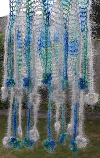 Bottom half of scarf hanging down outdoors showing fringe and first pattern repeat. The scarf is coloured with vertical stripes of variegated blue-greens and silver-greys. Each strand of fringe has a flower motif on its end.