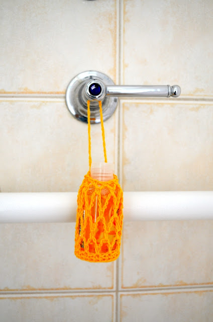 Front view of the shower gel hanger, hanging from a stainless steel shower tap and hanging in front of a white horizontal grab rail.