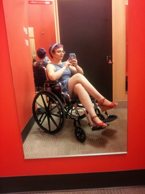 Young woman sitting in manual wheelchair, legs crossed, taking a selfie with a phone camera, viewed in a mirror
