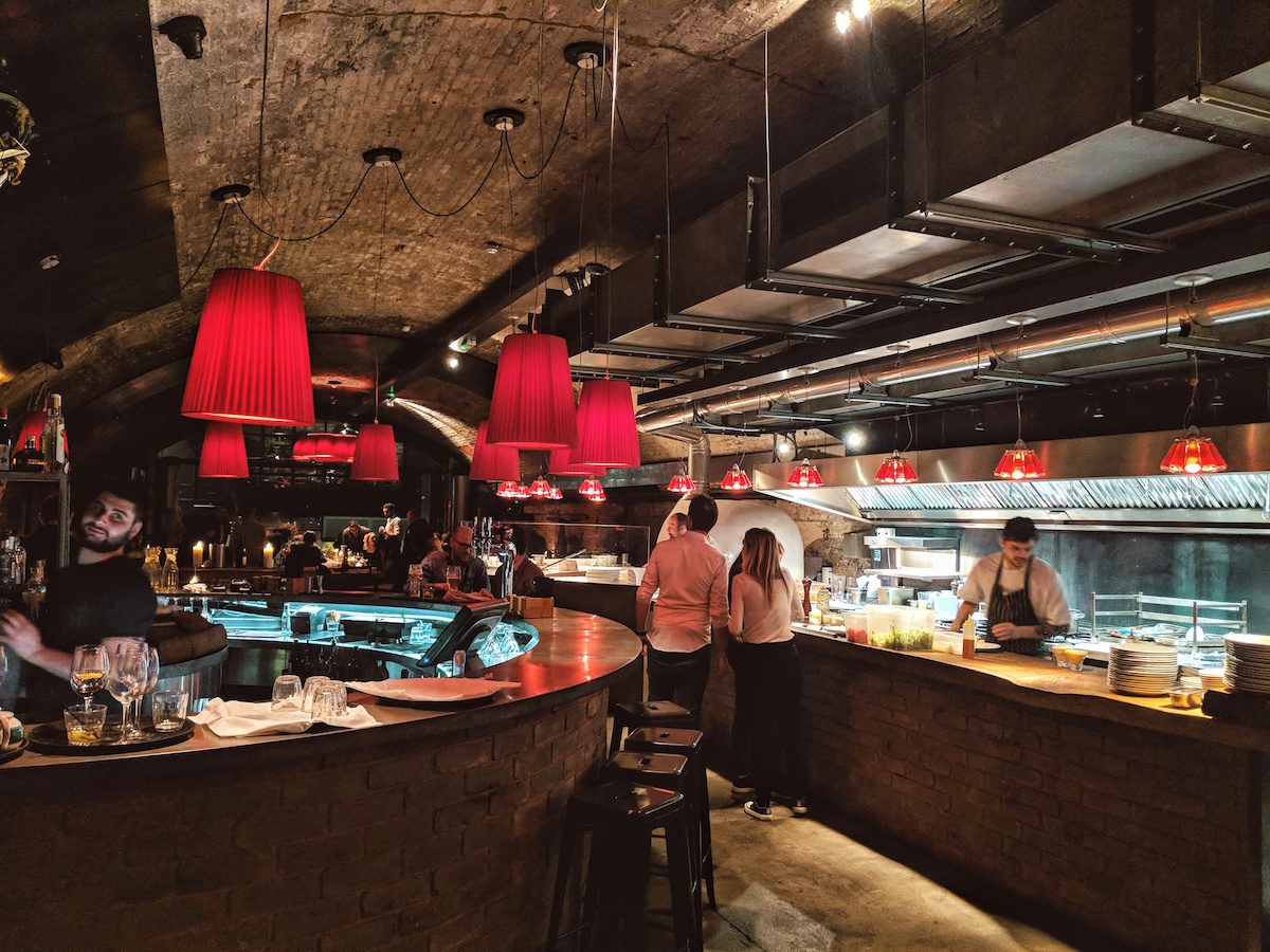 A well-stocked, half moon shaped bar sits opposite the open kitchen and Scandi red lampshades hang from the ceiling