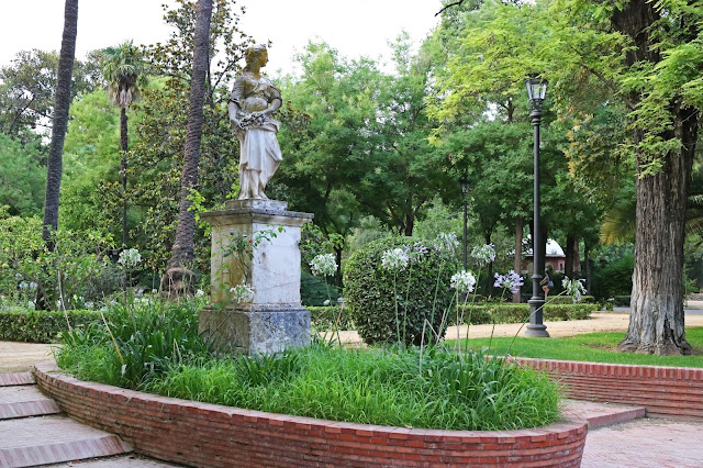 Una estatua femenina en un arriate en una placita de un parque con mucha vegetación.