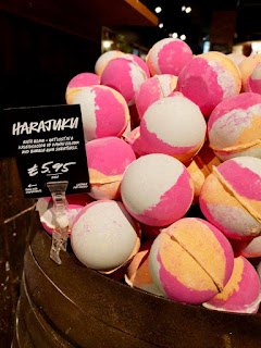 A selection of pink and yellow spherical bath bombs on a wide light brown rectangular shelf with a black rectangular board saying harajuku in white font on a bright background 