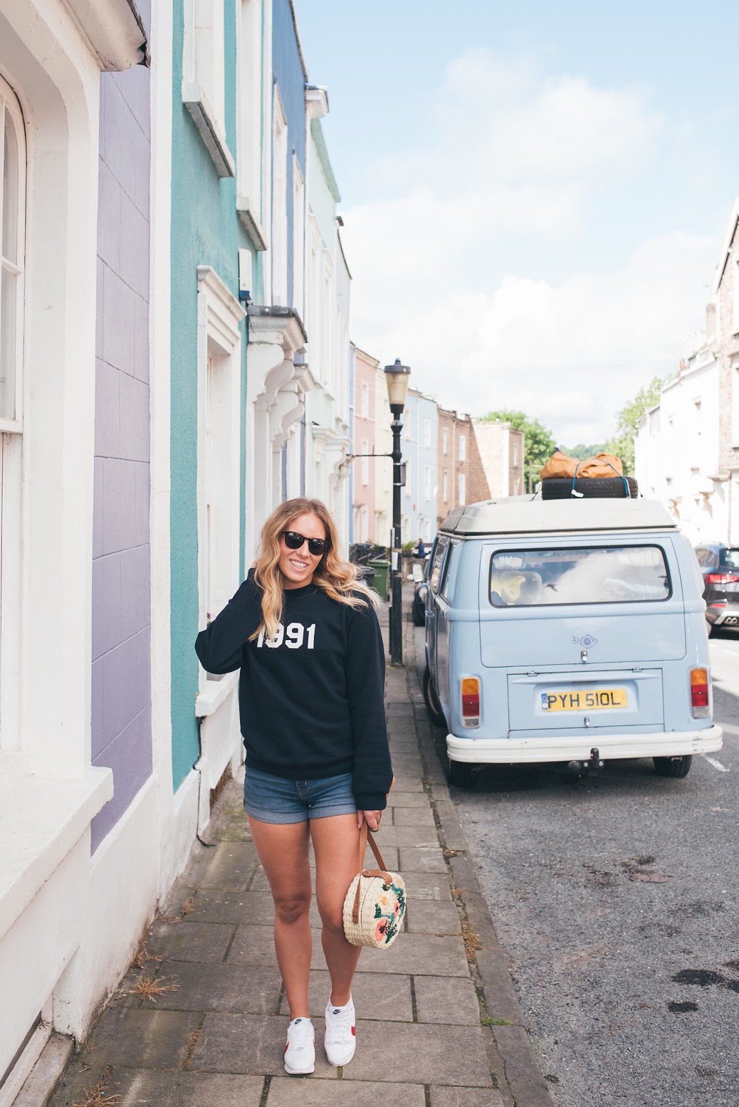 Rachel Emily in front of pastel coloured houses smiling with a pale blue VW Campervan