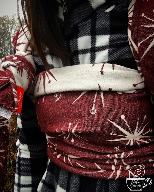 [Image of a of a woman’s torso focused on a maroon and ecru wool blend star burst patterned woven wrap carrier. Woman has on a black and white zip-up fleece jacket. The wrap carries a sleepy toddler on the woman’s back-though toddler isn’t visible in photo frame. The wrap is spread across the woman’s chest and double knotted at a shoulder; the top of the chest pass is folded down slightly so the reverse side is apparent. It’s a gray drizzly day but this woolie wrap turns up the cozy.]