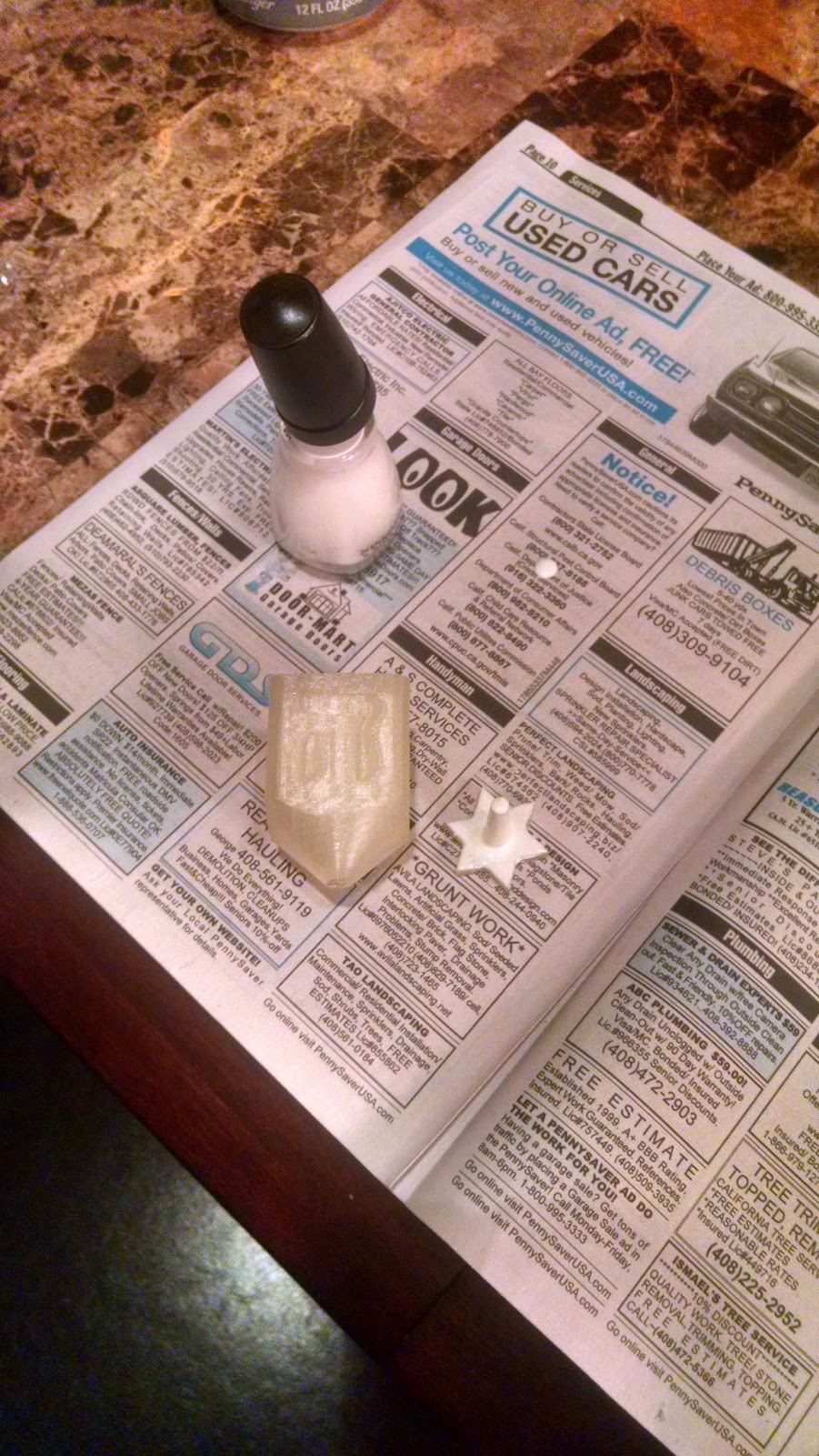 3D printed dreidel body and separate handle lying on newsprint. The handle is painted with white nail polish, the body is unpainted.