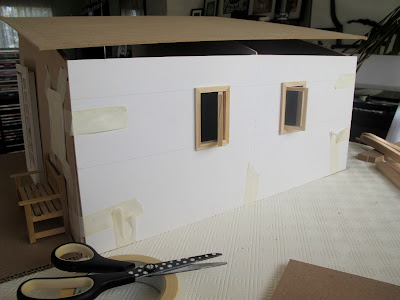 Rear view of a taped-together modern dolls' house miniature kit, with a cardboard wall and two small windows inserted.
