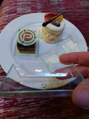 Plate with two small cakes on it. One is on a clear plastic tray. In the foreground is a hand holding up another of the trays.
