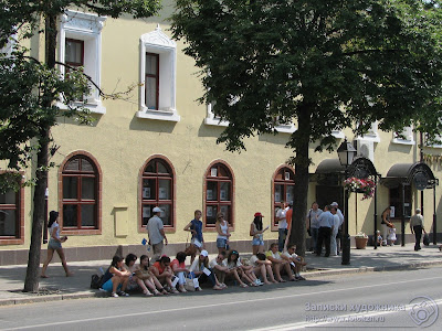 Зрители сидят в тени деревьев на поребрике улицы Кремлевской
