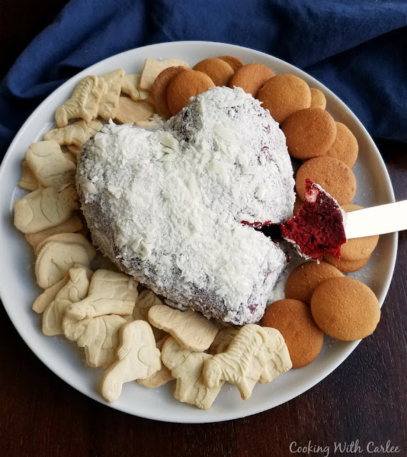 Luscious red velvet cake and cream cheese frosting flavors come together for a one of a kind dessert experience. Red velvet cheese balls are perfect for Valentine's Day, Christmas or just because! serving plate with heart shaped red velvet cheese ball coated in white chocolate shavings with nilla wafers and animal crackers for eating