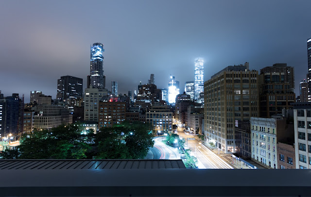 A nocturnal cityscape of Manhattan viewed from an elevated position, with the bright streaks of traffic lights painting ribbons of color along the streets. The dense array of buildings is punctuated by patches of greenery and illuminated by the soft glow of city lights, with skyscrapers rising majestically in the background, draped in a misty haze.