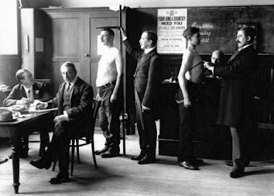 Black and white photo, WW1, a young man having his chest measured and another his height taken.  There are two men seated taking notes.  A recruiting poster is visible on the wall behind