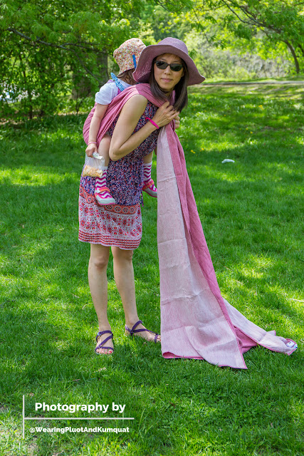 [Photograph by my husband. Image of me, a light tan skin Asian woman wearing sunglasses and a floppy purple sun hat, smiling at my husband who is taking our photograph. I have a preschooler on my back who is holding a bag of snacks and decided she needed to go up on my back to eat her snacks. I am using a pinkish reddish linen woven wrap and I have just made a seat for my child and I am holding on to the long beautiful freshly ironed tails of the wrap over my shoulders that are draped into the spring green grass.]