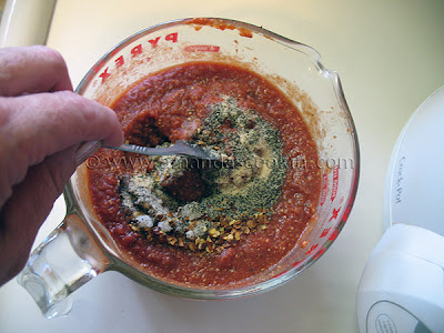 A photo of salt, pepper, garlic powder and pepper flakes being mixed into pizza sauce in a glass measuring cup.