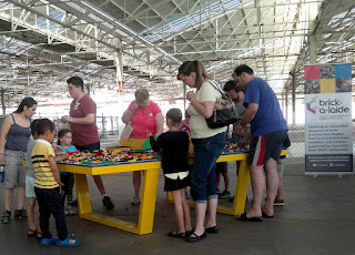 A large table similar to a table tennis table is covered with lego bricks and surrounded by children and adults who are playing with them.
