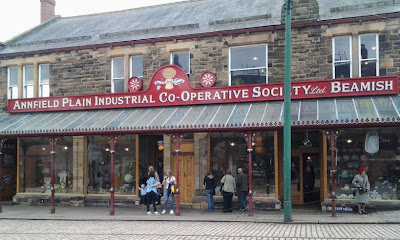A colour photo of the relocated Annfield Plain co-op store at Beamish, a two story stone building with a four plate glass windows and three entrances, clothing, food and hardware.  A glass canopy ties the shop fronts together and the road in front is cobbled with inlaid tracks for the trams.