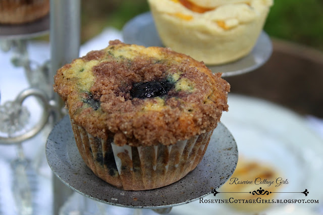Photo of a cupcake holder with blueberry muffin on one of the display places.  The muffin is in the paper liner and the top has a crumbly coating on top. Plump blueberries can be seen in the muffin.  This is a recipe by rosevinecottagegirls.com