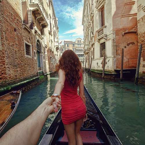 Fotografia da namorada de costas, puxando seu namorado pela mão esquerda, eles estão em uma canoa em uma das ruas alagadas de Viena, na Itália. se nota casas ou comércios em volta.