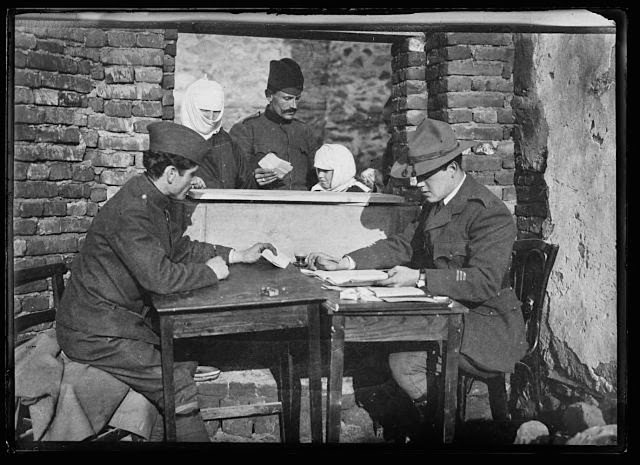From this little desk was done the paper work that distributed relief to many thousand Serbian refugees in the Monastir district. Captain Lanning Mc Farland, a Harvard student, was in charge of the American Red Cross work in this town. He operated a bakery, hospital, bathing establishment, warehouse and feeding station. He is shown at his desk examining the application of a Turkish woman for relief
