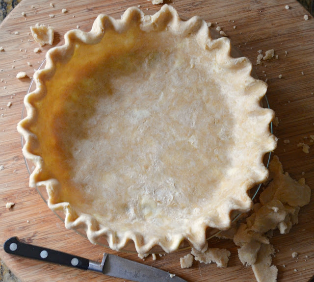 Uncooked flaky pastry dough in a glass pie plate shaped and the edge fluted on a bamboo cutting board with pieces of pie dough scattered around and a pairing knife laying on the board.