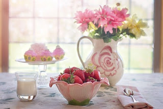 Uma bacia em forma de flor, cheia de morangos. Um copo de suco de morango. Um vaso de flores. Uma compota com doces de morango.