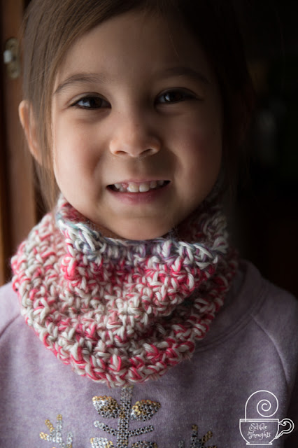 Image of a smiling child with twinkling eyes wearing a multi-colored crocheted cowl over a lavender sweatshirt featuring a silver sequin snowflake.