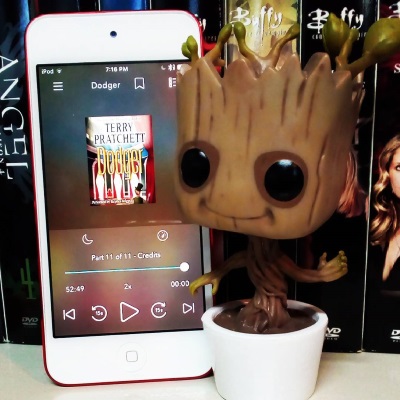 A large-headed Funko Pop bobblehead of Dancing Groot stands next to a white iPod with Dodger's cover on its screen. The cover image is very small, but the viewer can barely make out a stage draped with red velvet panels.