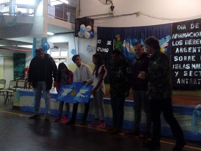 se observa a alumnos sosteniendo un cuadro pintado por ellos de las islas malvinas, de pie junto a los ex combatientes