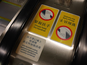 'Always hold the hand rail' and 'Anti-bacterial coating applied to handrail with addition disinfection regularly carried out' signs on an MTR escalator in Hong Kong