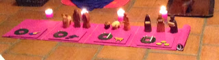 This photo has been cropped, so it is short and long, showing just the Godly Play Advent materials - - a long strip of purple cloth, with four plaques on it (exactly matching the purple of the cloth), various figures from a wooden nativity set, and four lit candles behind it.