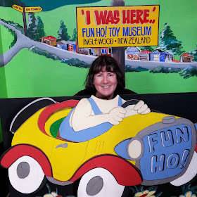Woman posing in a cardboard car in front of a sign saying 'I was here...Fun Ho! Toy Museum'