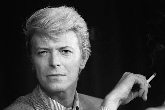 image en noir et blanc de David Bowie, cet artiste est décédé d'un cancer le 10 janvier 2016. la photo le montre fumant une cigarette et fixant l'objectif