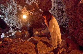 Woman in prayer at back entrance to cave facing water