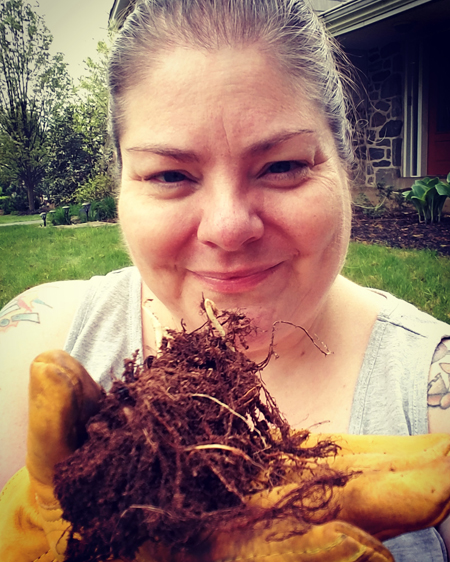 image of me in my front yard, holding up an earthy bud in yellow gardening gloves; I'm wearing a grey tank top and my contacts, with my hair pulled back