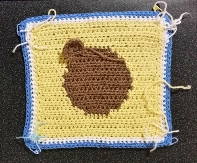 A yellow square crocheted in rows with a white and blue border crocheted in two rounds. A tan intarsia circle is in the middle. One ear is stitched to what looks like the top-left of the circle.