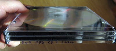[Image: CD jewel cases with text labels hand-written on the backs but mostly invisible because of the lack of contrast between the black ink and the black plastic showing through the transparent case.]