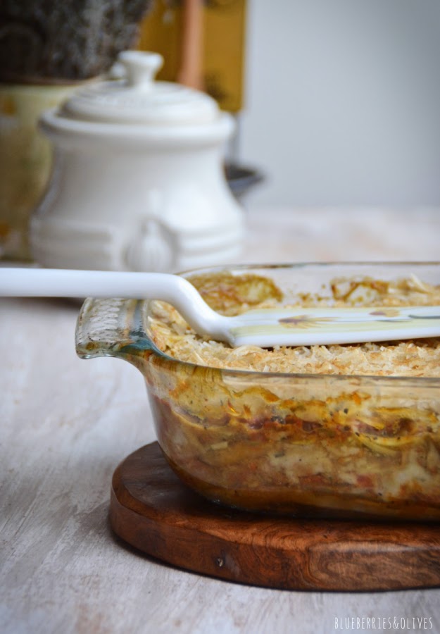 Lasaña de berenjenas en fuente de cristal sobre tabla de madera, con ingredientes de fondo, hierbas aromáticas, laurel seco, molinillo pimienta, espátula vintage para servir