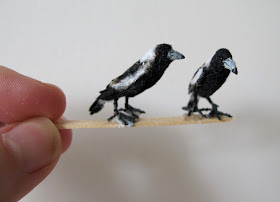 Two one-twelfth scale magpies perched on a piece of wood held by a hand.