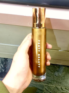 A white hand holding a large cylindrical glass bottle with Fenty Beauty written in block white small font on the front of it with a light brown sparkly liquid on the insde on a light background. 