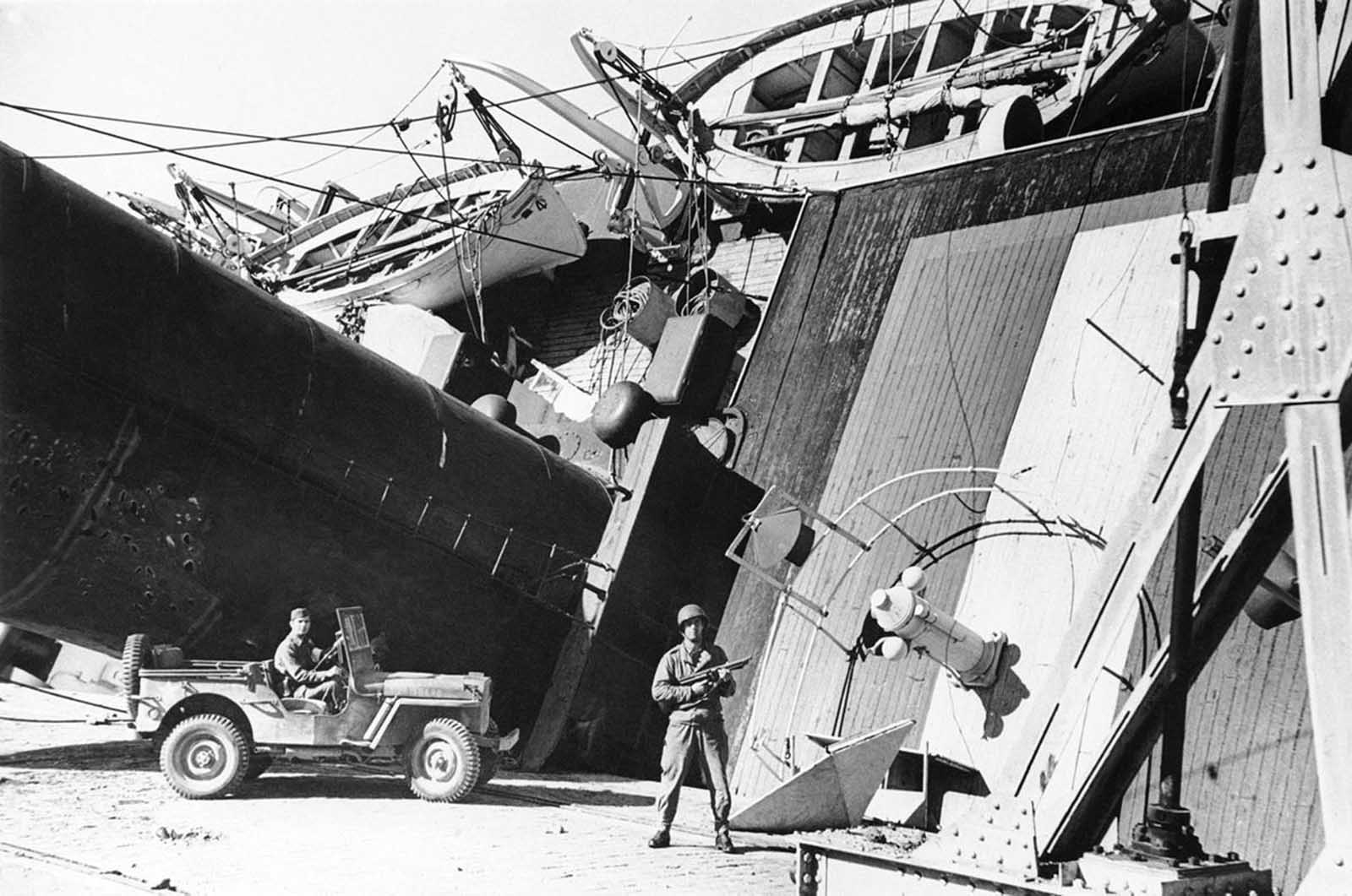 A U.S. army soldier with a sub-machine gun and another in a jeep guard the looming S. S. Partos which was damaged and had capsized against the dock when the Allies landed at the North African port, in 1942.