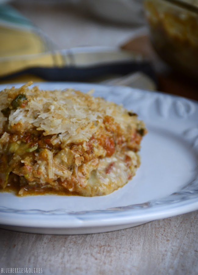 Trozo de lasaña cortado sobre plato blanco, Lasaña de berenjenas en fuente de cristal sobre tabla de madera, con ingredientes de fondo, hierbas aromáticas, laurel seco, molinillo pimienta