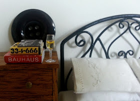 One-twelfth scale modern miniature scene of a black iron bed with white embroidered bedding next to a wooden chest of drawers with a stack of design books and a glass of sparking wine on it.