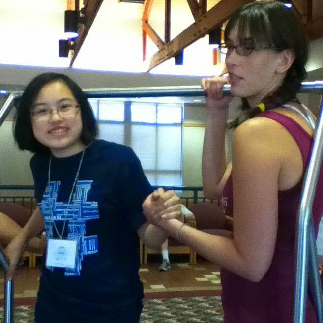 Image is 2 medium to fair skinned female presenting people, both with dark hair, dark eyes, and glasses. The person on the left has short black hair, is visibly of Chinese descent, and is wearing a blue t shirt. The person on the right has long very dark brown hair, is ethnically ambiguous, and is wearing a burgandy tank top. They are standing on a hotel luggage cart.