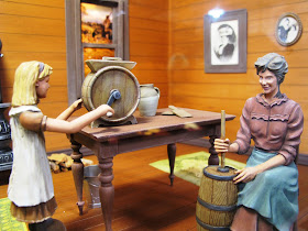 Model of a 19th-century woman and child inside a house, churning butter.