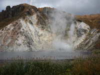 Larger sulfurous hot pond near noboribestu-onsen emitting some steam with white hill in background with trees on the top