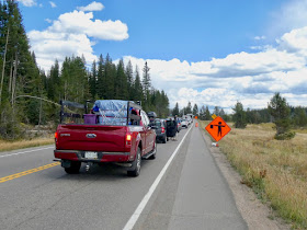 Trucks and cars lined up on the road, road construction sign on the right, forested land on the left