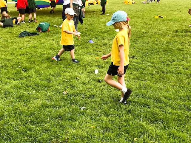 A young girl in yellow t-shirt, black shorts and blue hat is walking with good posture in the egg and spoon race. The egg has just fallen off the spoon.