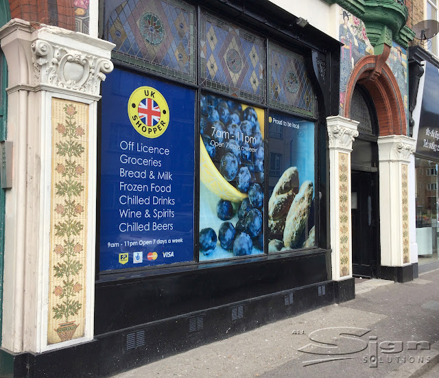 An old building with wonderful japanese painted figures on the door opening. The stained glass windows reflect the light. In the large windows, printed vinyl stickers have UK Shopper logo, Off Licence, Groceries, Bread & Milk, Frozen Food, Chilled Drinks, Wine & Spirits, Chilled Beers. Open seven days a week. Proud to be local shop.