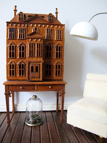 Modern dolls' house miniature scene with a wooden dolls' house mounted on a desk next to a white slipper chair, an arc lamp and with a cloche on the floor underneath it.