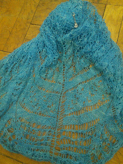 A blue lace shawl live on the needles  The shawl is spread out to show various lace panels and patterns.  A stitch marker with a shell charm is clipped into the knitting. 